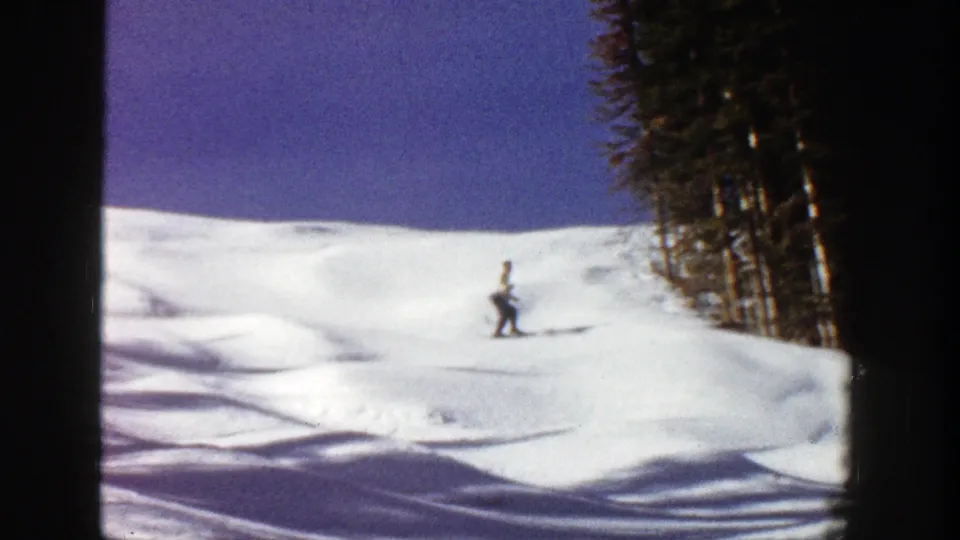 A distant skier with poles crosses open snow in the Aspen mountain setting, framed by trees and blue sky.