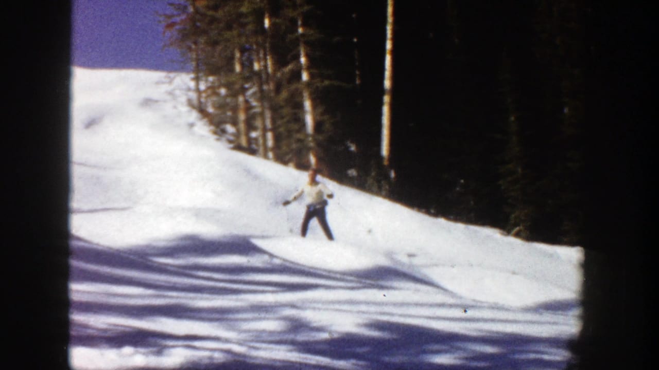 A skier in light jacket and dark pants descends a sunny Aspen, Colorado slope in 1961, framed by evergreen trees.