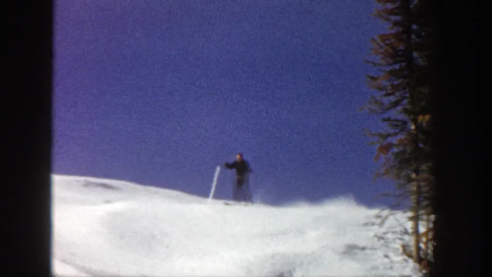 A skier in dark winter clothing drops down the snowy Aspen slope, with powder spray and evergreen trees at the edge of frame.