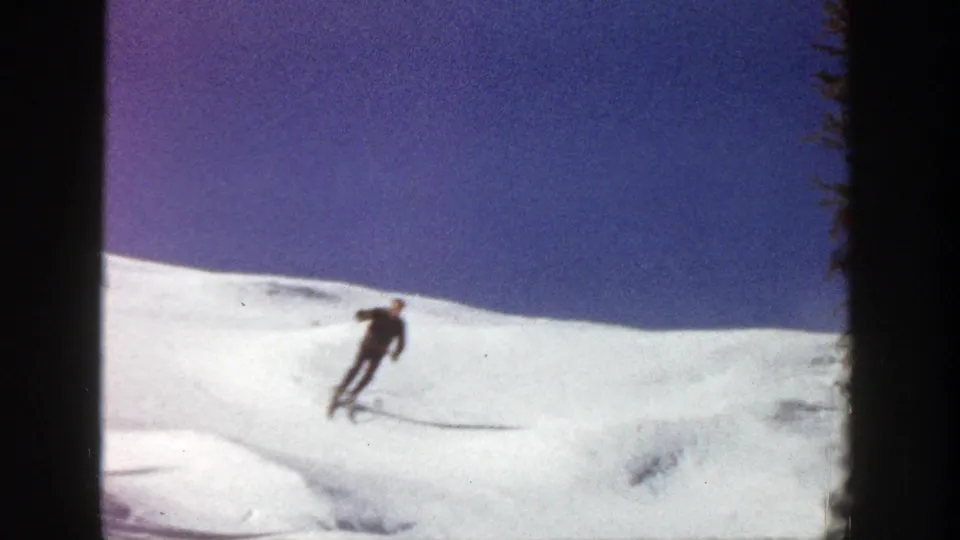 The skier carves down the Aspen snow slope in a distant 1961 home-movie view, leaning into the turn against a blue sky.