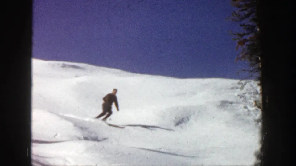 A distant skier moves across a white Aspen snow slope in 1961 home-movie footage, with trees at the edge of the run.
