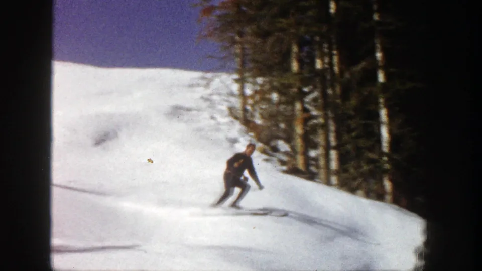 A skier in dark clothing carves across a sunlit Aspen slope, with dense evergreens visible along the right side.
