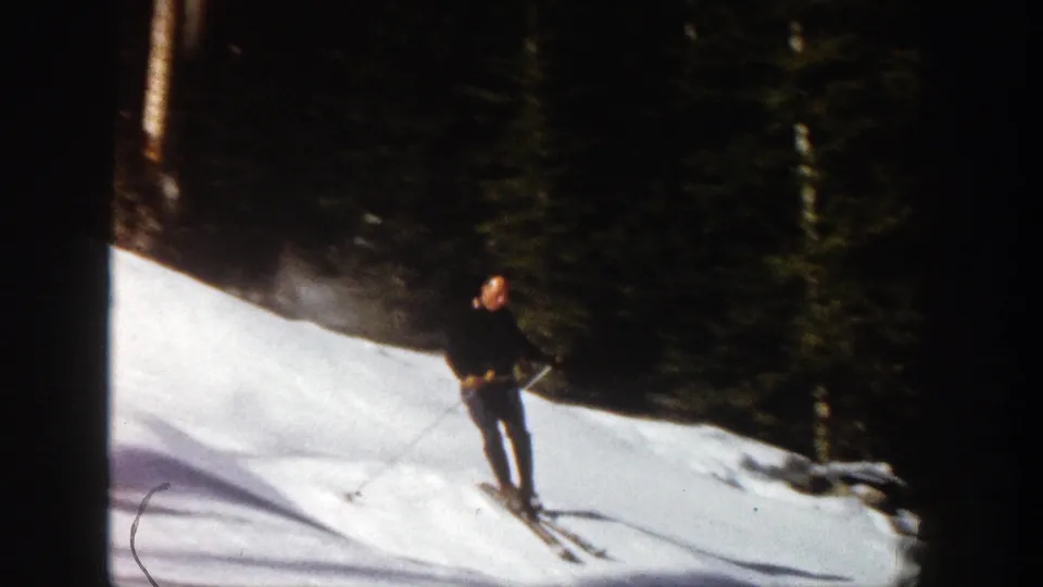 The skier continues downhill in 1961 Aspen footage, framed against snow, tree shadows, and a dark evergreen background.