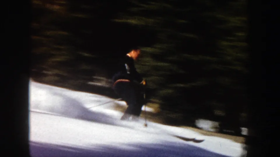 A skier in dark clothing travels along the snow near a shadowed evergreen treeline in vintage Aspen home-movie footage.
