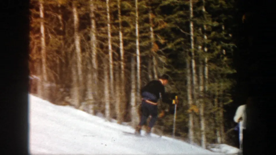 A dark-clothed skier crosses a snowy forested slope in Aspen, with another skier partly visible at the right edge.