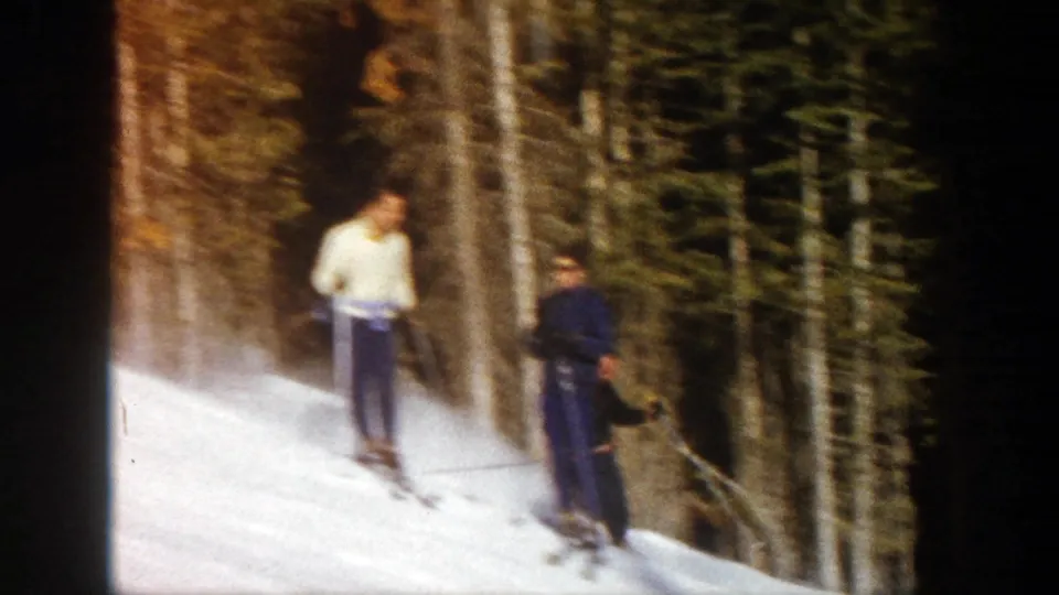 Two skiers, one in a light top and one in darker clothing, pause or descend slowly beside trees in 1961 Aspen ski footage.