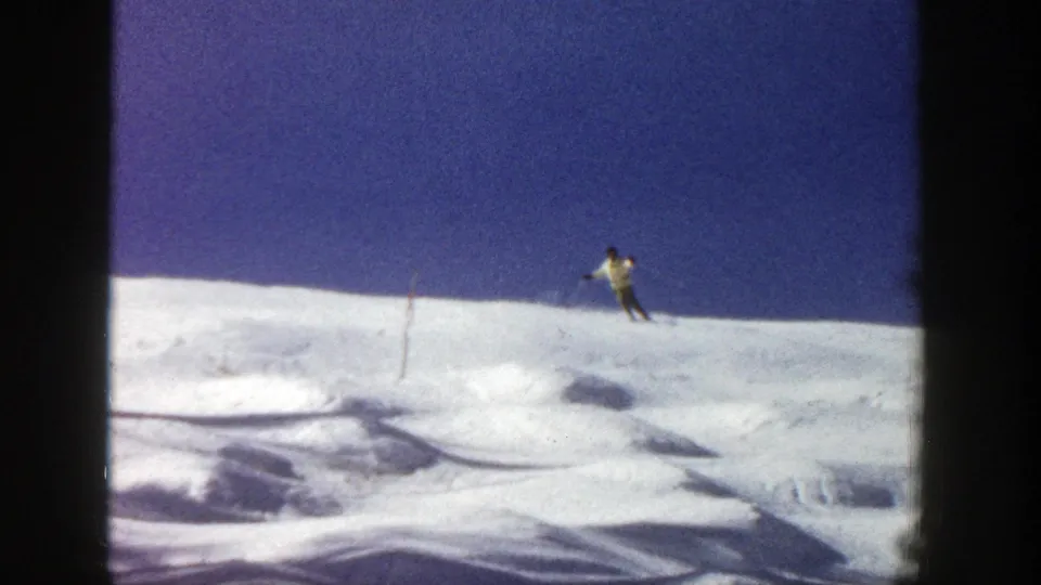 A distant skier moves across an open Aspen snowfield beneath a clear blue sky, with ski tracks and shadows visible in the foreground.