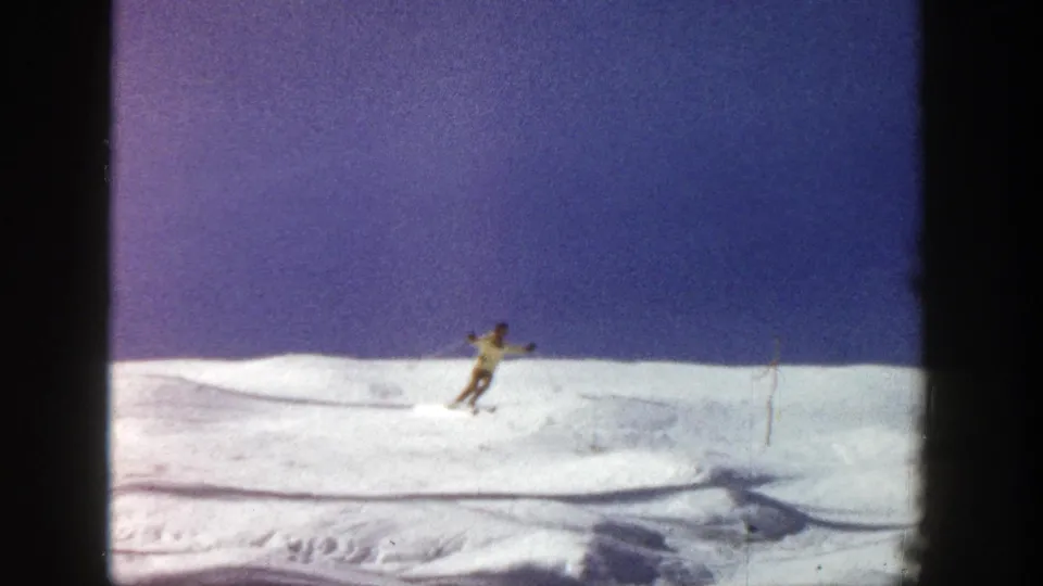 A skier descends diagonally through sunlit snow in 1961 Aspen footage, framed at a distance in a home-movie view.