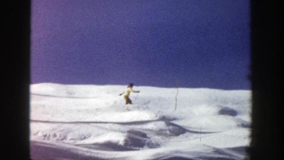 Distant 1961 Aspen frame of a skier gliding across a broad snow-covered hill, with ski tracks and wind-shaped snow visible.