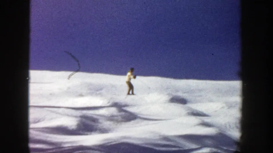 A lone skier in Aspen, Colorado, 1961, stands or glides across an uneven snowy slope under a deep blue sky.