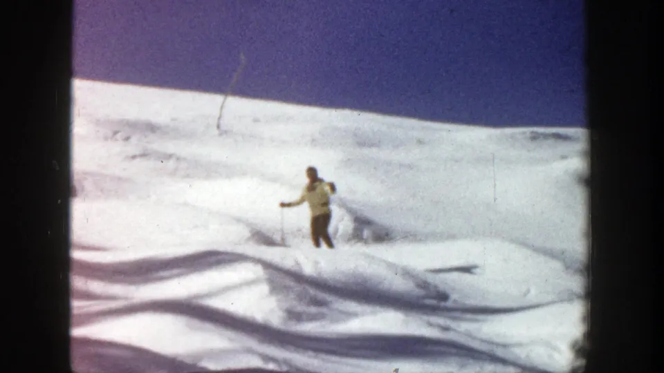 Adult skier continues downhill on an undulating Aspen snowfield, framed from a distance in 8mm home-movie footage.