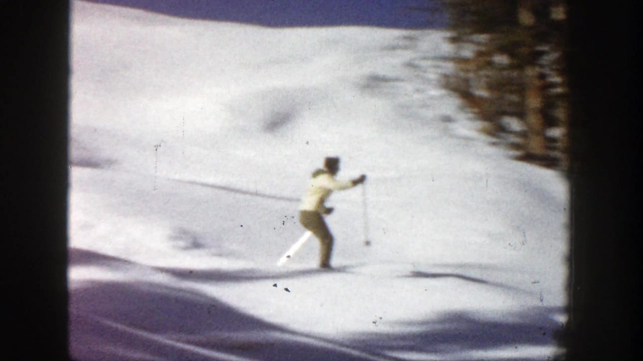 Woman in a yellow jacket skis across a snowy slope with evergreen trees behind her.