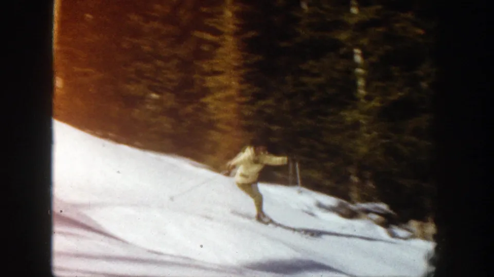 The adult skier in yellow winter clothing descends a snowy Aspen slope, with dark evergreens and warm 8mm color in the background.
