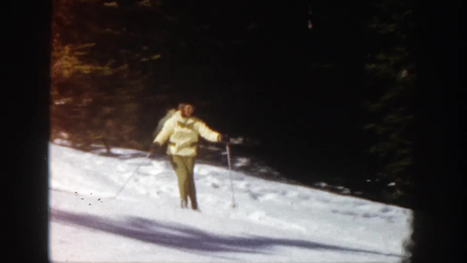 An adult woman skier in yellow winter clothing faces the camera on a snowy Aspen slope, holding ski poles near the wooded edge.