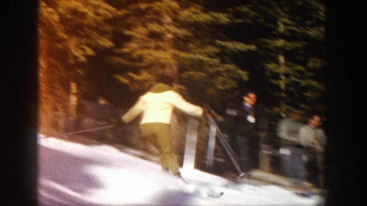 Skier in a light jacket descends a snowy slope near trees and onlookers.
