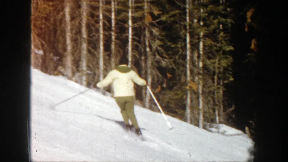 1961 Aspen frame with a skier seen from behind, using poles while moving across a bright snow slope beside tall evergreens.