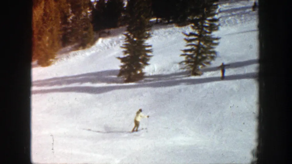 Wide 1961 Aspen ski-slope view showing a skier lower left, evergreen trees, long shadows, and small distant skiers on the snow.