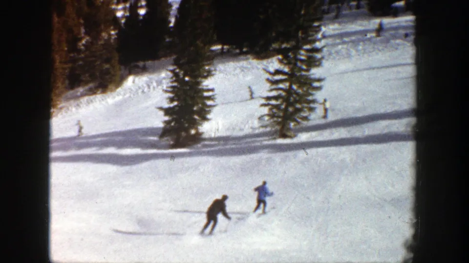 Two distant skiers cross a sunlit Aspen slope in 1961, with evergreen trees and long shadows behind them.