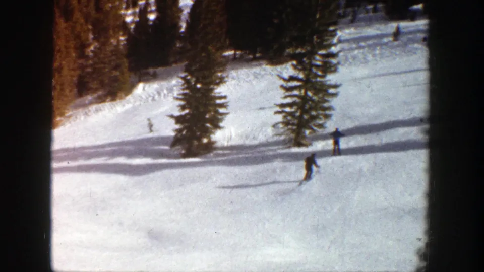 A 1961 Aspen ski slope appears with several distant figures, evergreens, and bright snow in soft 8mm color.