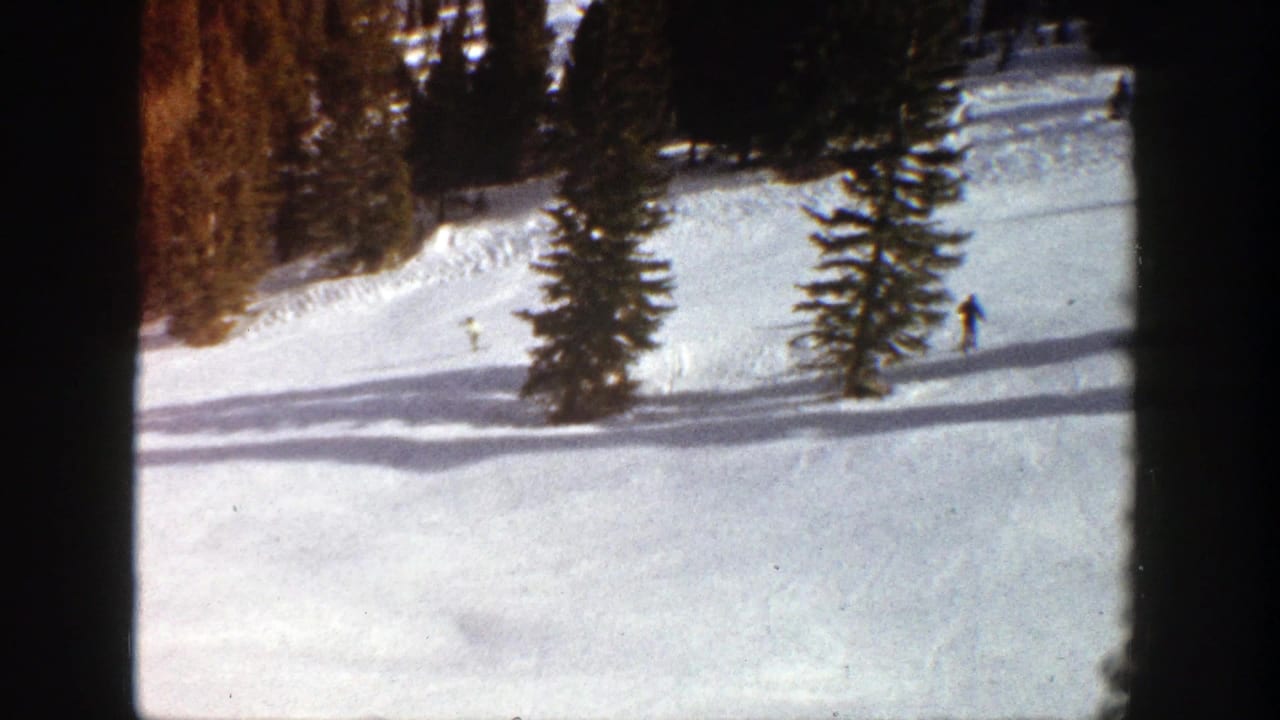 In 1961 Aspen, Colorado, distant skiers move across a bright snowy slope with evergreen trees and long shadows in an 8mm home-movie frame.