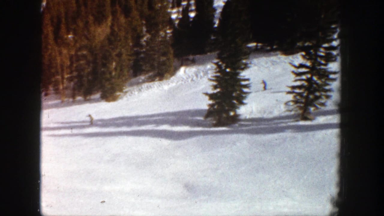 Distant skiers appear on a sunlit Aspen snow slope in 1961, framed by evergreen trees and dark 8mm film borders.