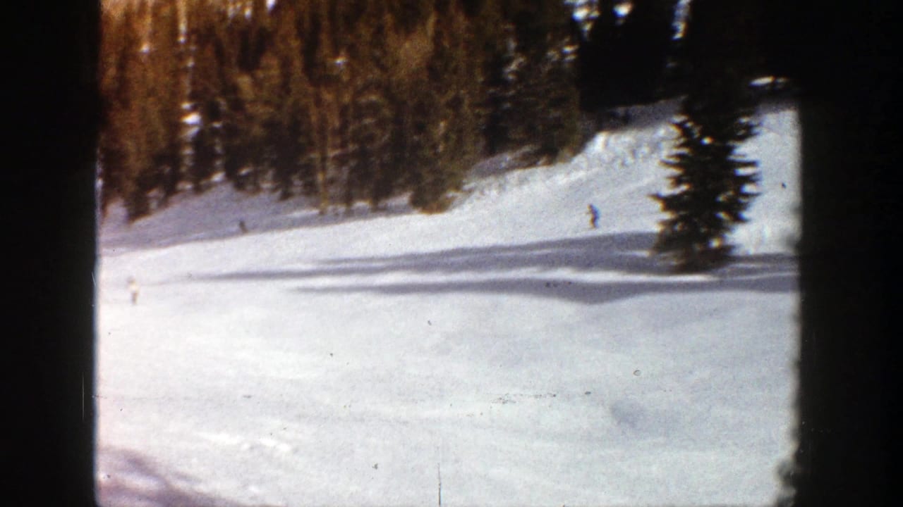 A snowy ski slope with distant skiers and evergreen trees in soft 8mm color.