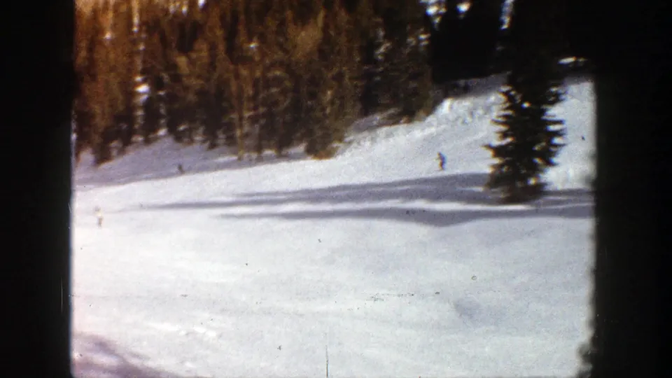A 1961 Aspen ski slope is seen from a fixed home-movie viewpoint, with distant skiers crossing the snow near tall evergreens.