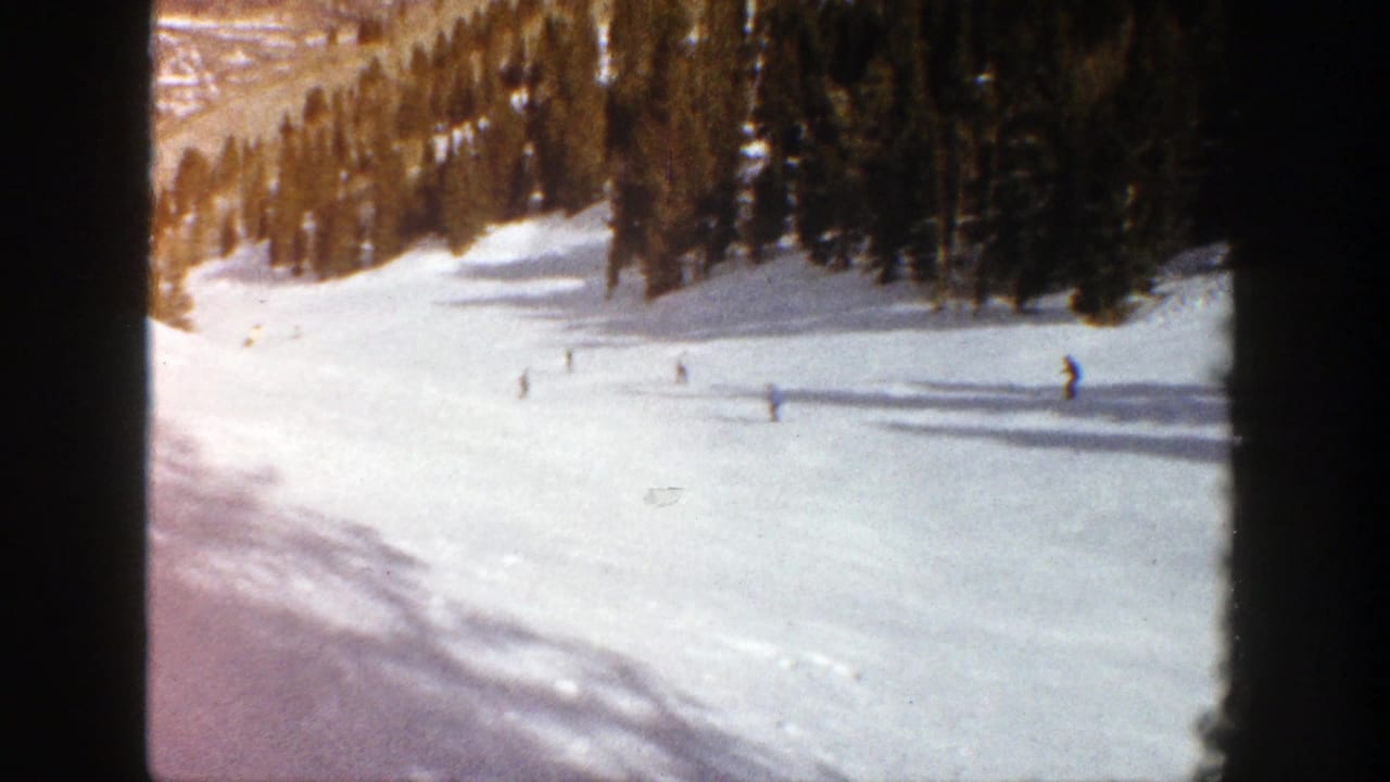 1961 Aspen home-movie frame showing small skiers on a sunlit snow-covered slope with dark evergreen trees and long shadows.