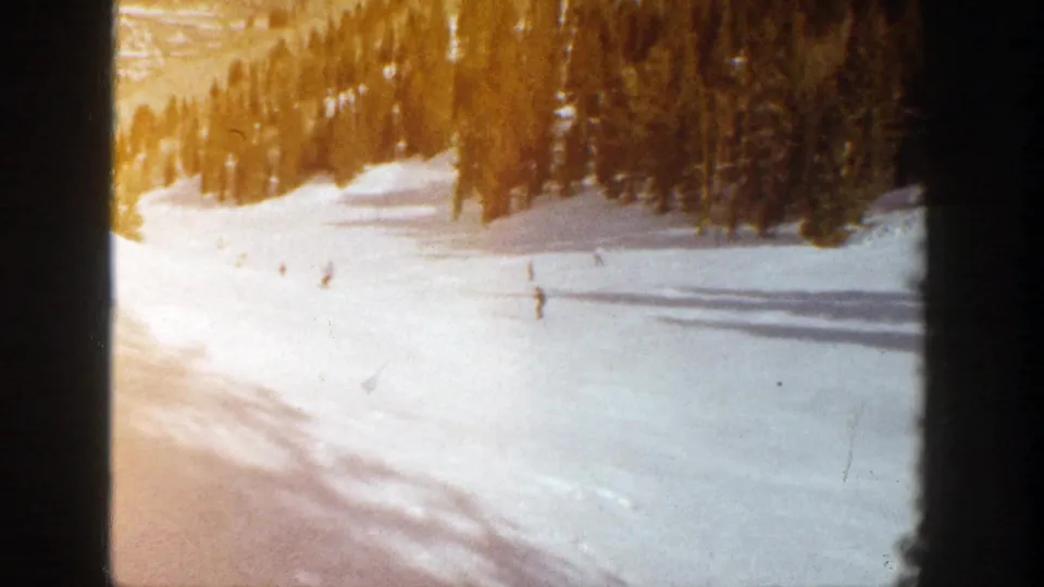 1961 Aspen 8mm frame with distant skiers spaced across a sunlit ski slope, tree line at right, and warm film color.
