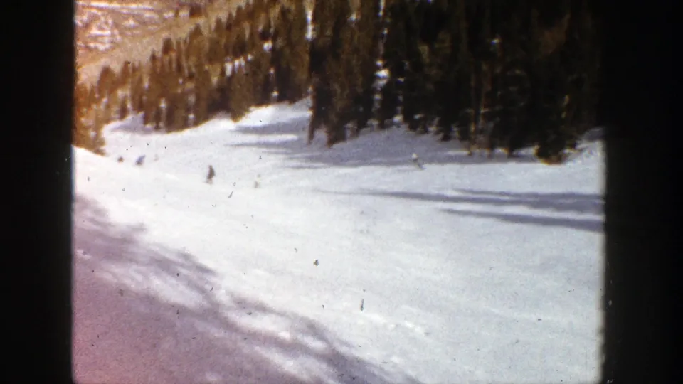 Distant 1961 Aspen skiing footage shows one darker figure lower on the snow with additional skiers near the tree-lined upper slope.