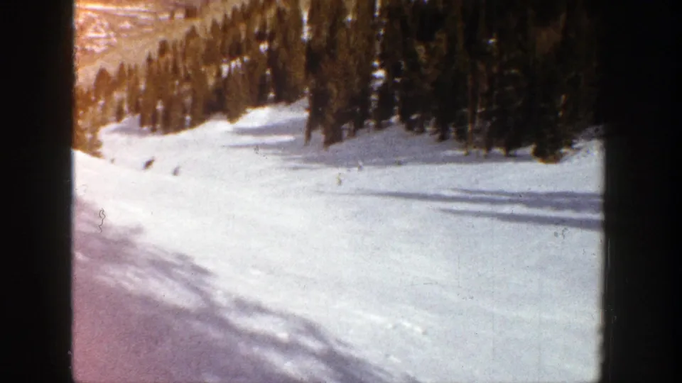 End frame from 1961 Aspen home-movie skiing footage, with distant skiers on a broad snowfield and dense evergreens along the run.