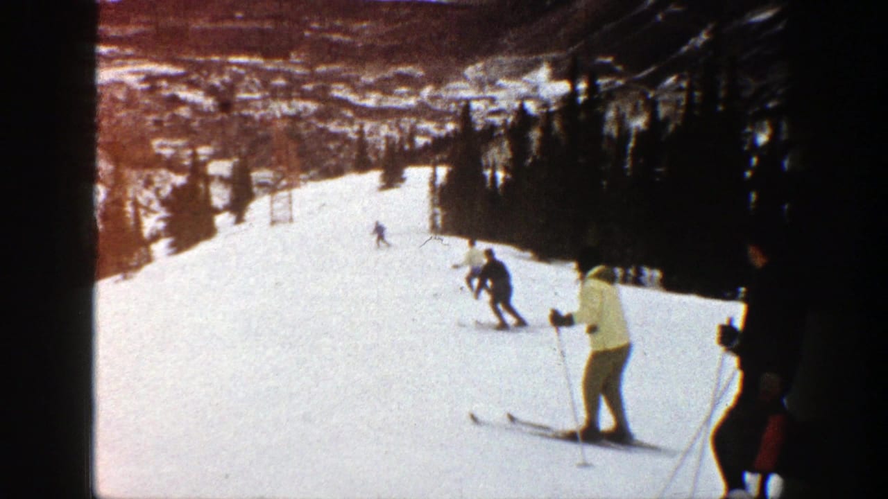 1961 Aspen home-movie frame showing skiers moving down a snowy run with evergreen trees and distant hills behind them.
