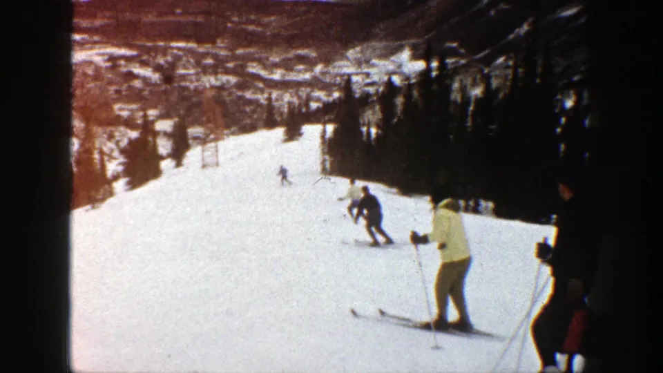 1961 Aspen home-movie frame showing skiers moving down a snowy run with evergreen trees and distant hills behind them.