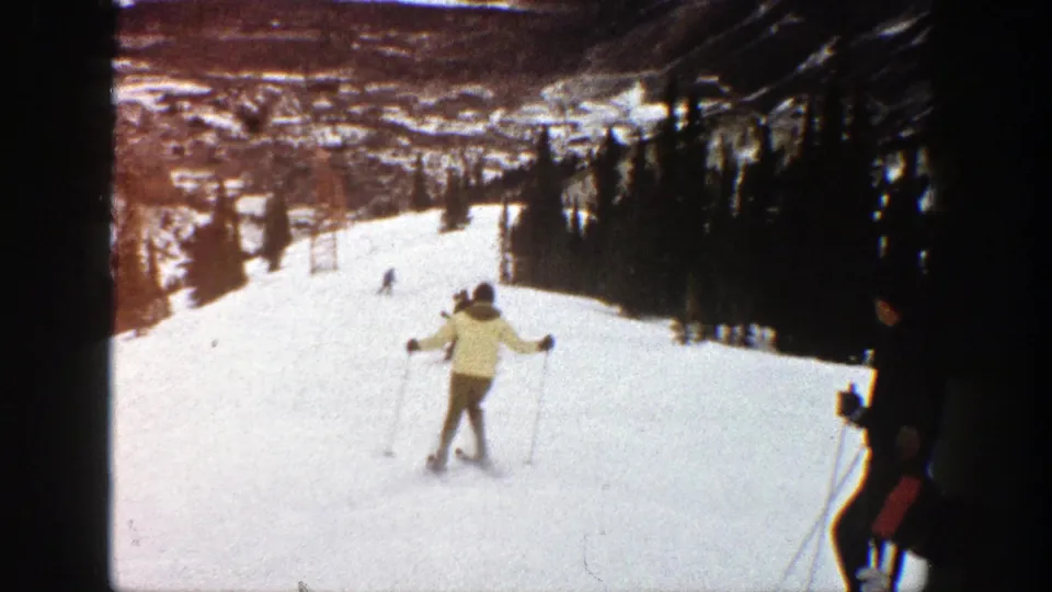 A skier in a light coat and dark pants descends an Aspen ski run, with other skiers and forested slopes in the background.