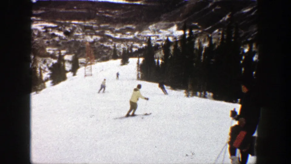 Vintage 8mm frame from Aspen showing a skier crossing the foreground of a snowy run with several distant skiers near the tree line.