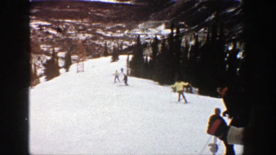Wide 1961 Aspen ski-slope view with several skiers on snow, a dark evergreen border, and a figure partly visible at the right edge.