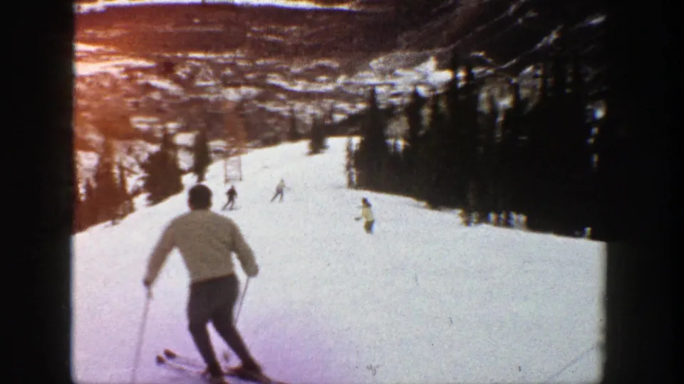 1961 Aspen frame with a foreground skier in a light jacket descending toward scattered skiers on a tree-lined snowy run.