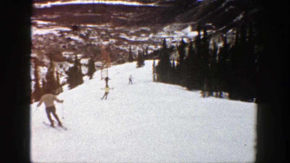 1961 Aspen home-movie frame showing a skier at lower left and several distant skiers descending a broad mountain slope.