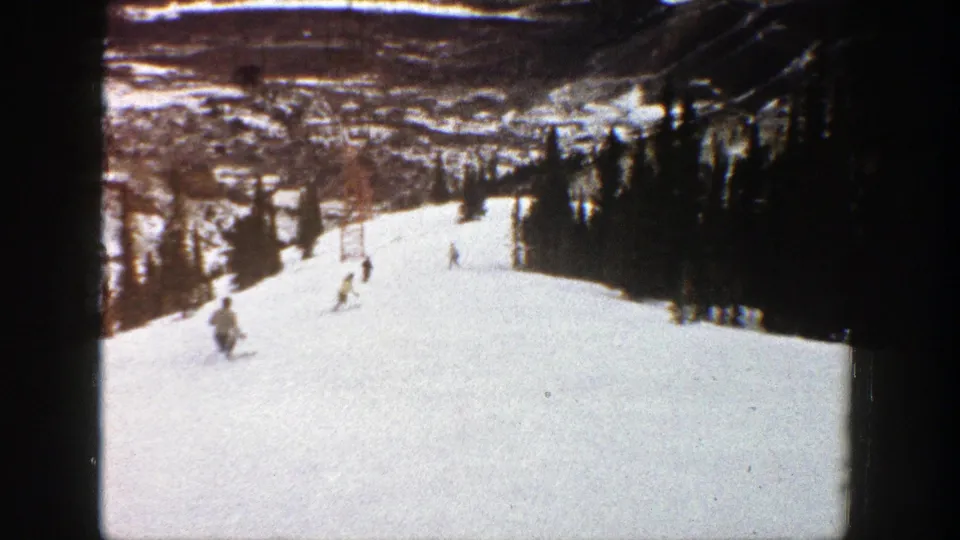 1961 Aspen frame showing small skiers spread across a snowy run, with evergreens and a valley landscape beyond.