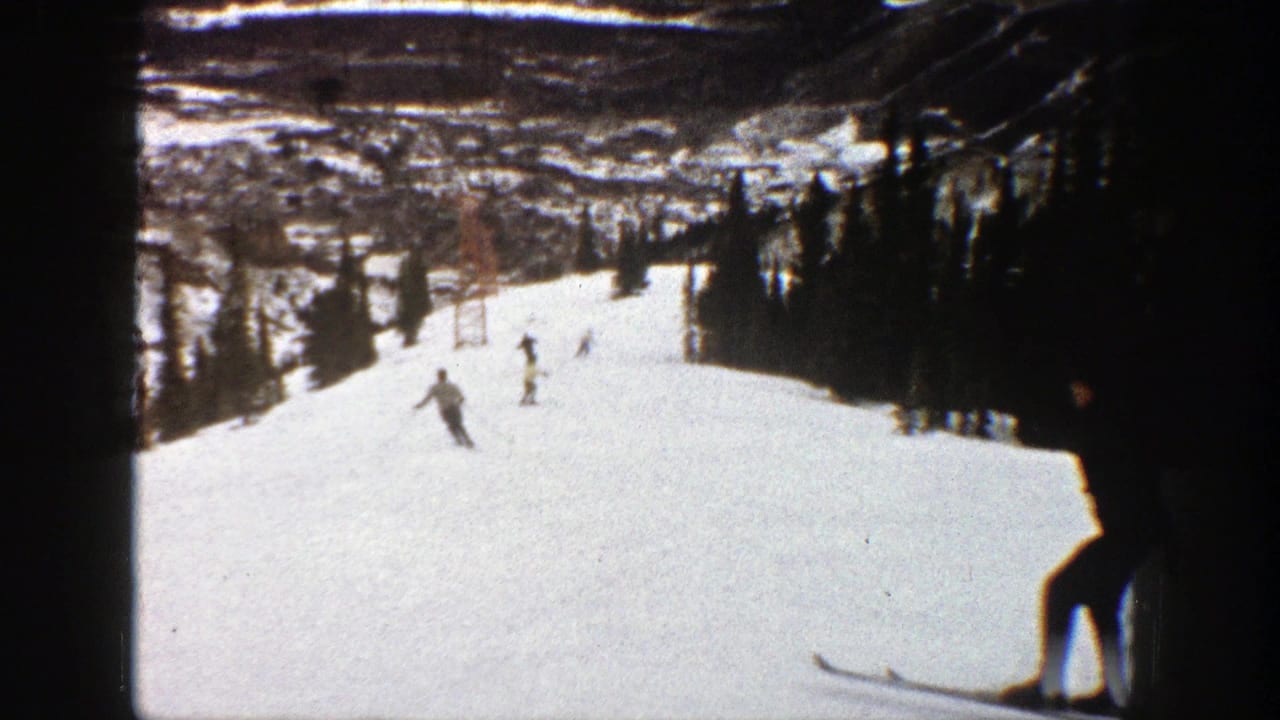 1961 Aspen home-movie frame showing several skiers moving downhill on a wide snowy run with trees at the sides.