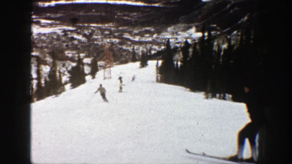 1961 Aspen home-movie frame showing several skiers moving downhill on a wide snowy run with trees at the sides.