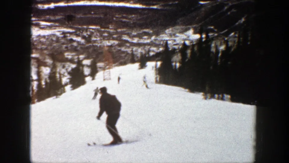 A skier in dark winter clothing glides across the foreground of a 1961 Aspen ski-slope home movie.