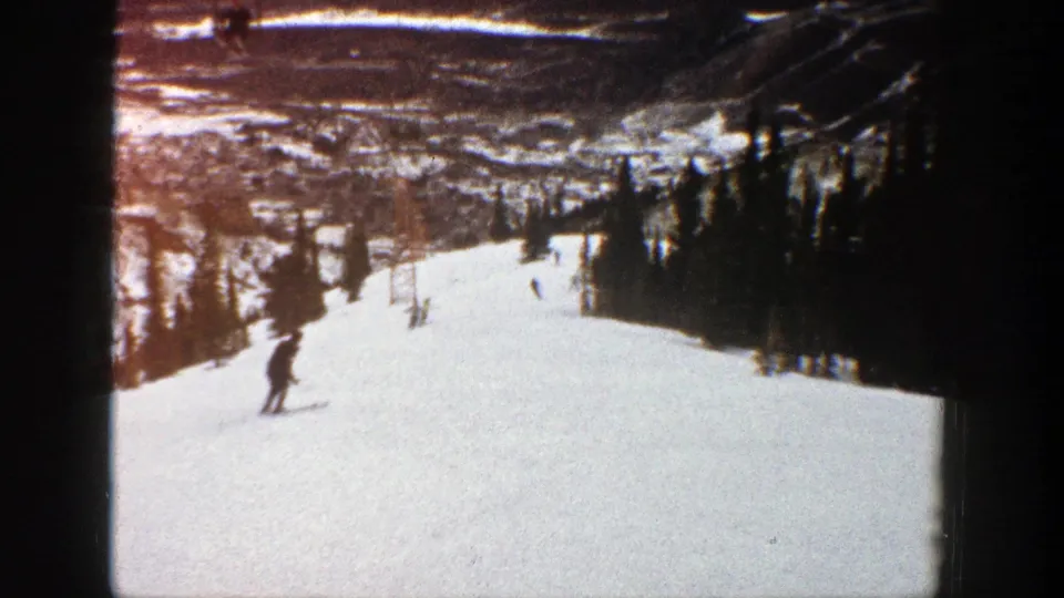 A skier crosses the lower slope while other distant skiers descend an Aspen run bordered by evergreens.