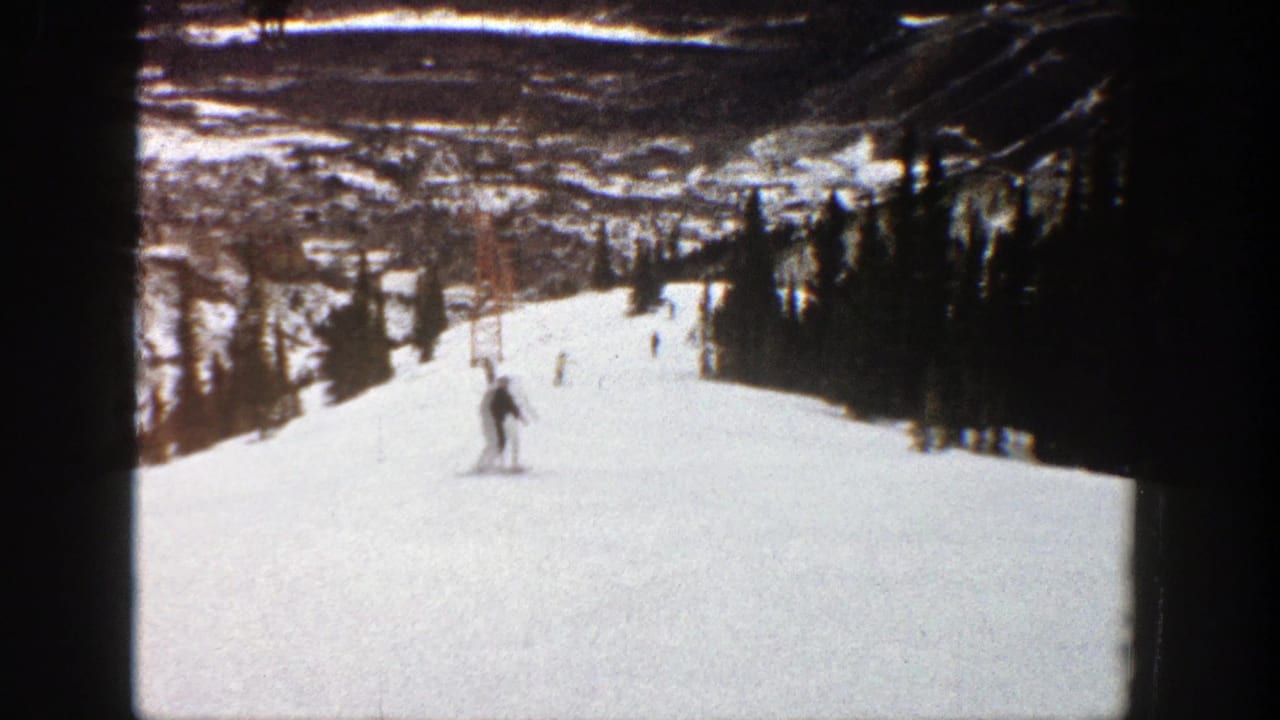 1961 Aspen home-movie frame showing a skier on a broad snowy run, with evergreen trees and distant mountain terrain visible.