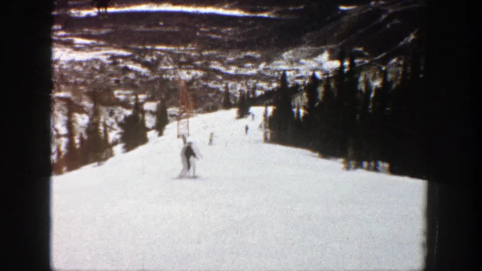 1961 Aspen home-movie frame showing a skier on a broad snowy run, with evergreen trees and distant mountain terrain visible.