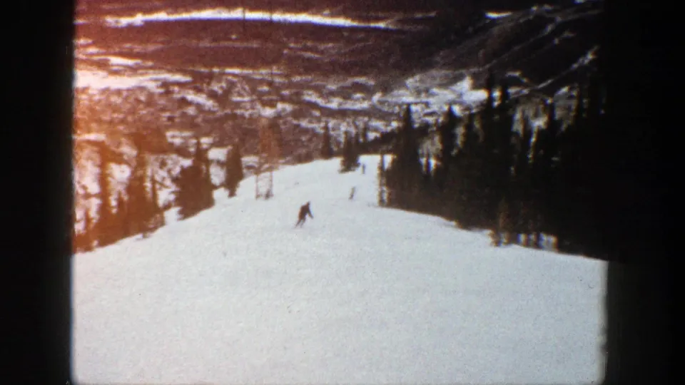 Faded 8mm frame from Aspen in 1961 showing a skier carving across a snowy slope beneath distant mountain scenery.