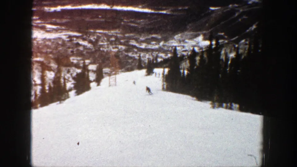 1961 Aspen home-movie still with a skier near the tree-lined side of a bright resort slope and distant hills behind.