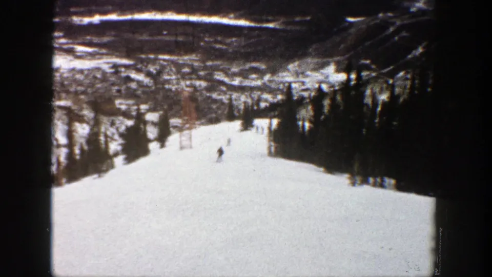 1961 Aspen home-movie frame showing skiers on a broad snowy run bordered by evergreens with a distant mountain valley beyond.