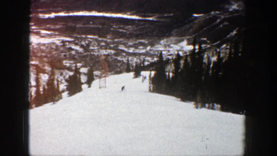 1961 Aspen ski footage frame with a broad white slope, small downhill figures, evergreens, and a hazy valley landscape in the background.