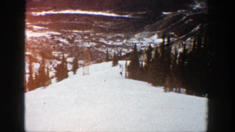 1961 Aspen frame showing skiers partway down a snowy mountain run, with dark evergreens at the side and valley terrain beyond.